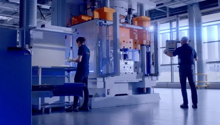 Twee medewerkers werken in een lichte industriële hal bij een grote persinstallatie, waarbij één operator onderdelen positioneert en de ander de machine bedient via een bedieningspaneel, in een strak georganiseerde en technisch geavanceerde productieomgeving van Bosch in Tilburg.