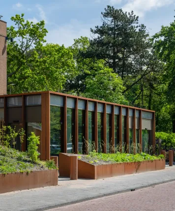Gevel van een moderne glazen uitbreiding met roestkleurige staalconstructie, omgeven door groene beplanting en volwassen bomen.