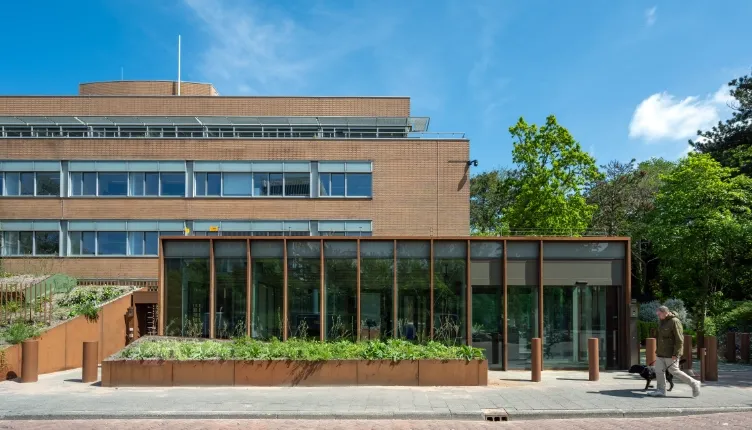 Vooraanzicht van een modern kantoorgebouw met een lage glazen aanbouw, grote raampartijen, plantenbakken en bomen langs de straat op een zonnige dag.