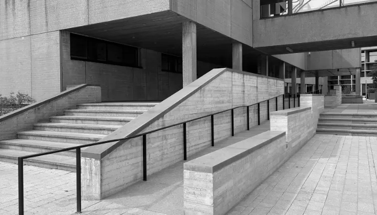 Toegankelijke entree van een modern gebouw met brede betonnen trappen, een centrale hellingbaan en metalen leuningen in een minimalistische architectuurstijl.