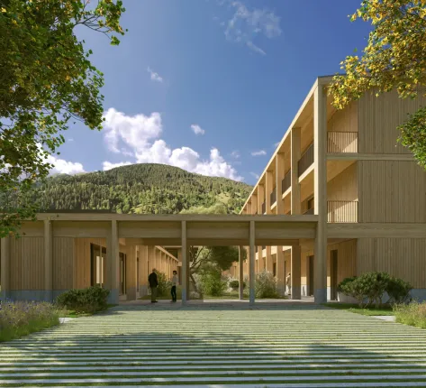 Houten woonzorggebouw in een bergachtig landschap met overdekte entree, groene binnentuin en zicht op beboste hellingen onder een heldere blauwe lucht.